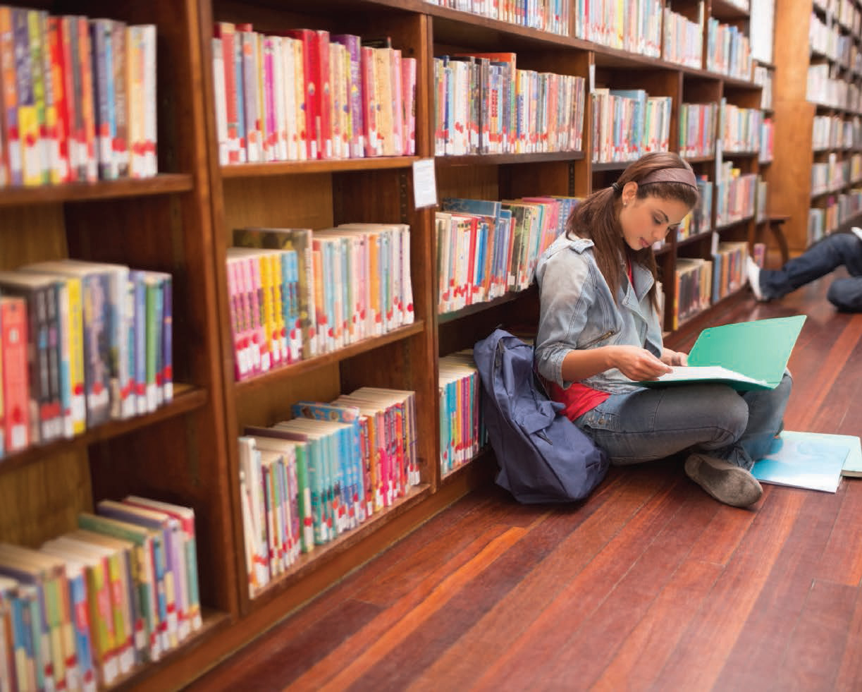 A student works on homework in the library sitting against a bookshelf