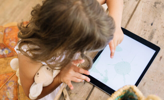 A little girl draws on her iPad while holding a stuffed animal