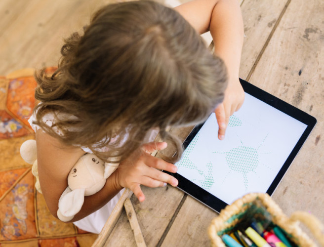 A little girl draws on her iPad while holding a stuffed animal