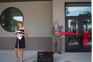 Executive Director Jackie Collins speaks while Principal Anthony Haskett holds the red ribbon for the opening