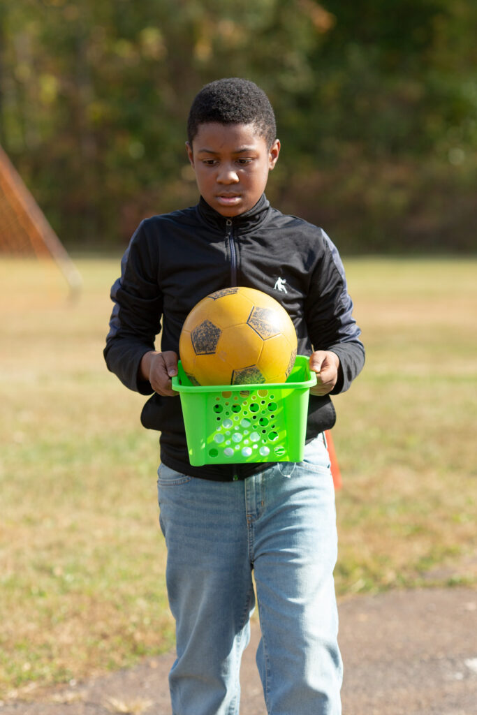 A kid carries a soccer ball outside without a face mask on