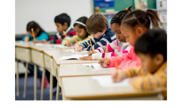 Elementary school students seated at classroom desks, writing in notebooks and completing schoolwork.