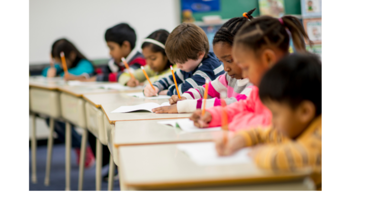 Elementary school students seated at classroom desks, writing in notebooks and completing schoolwork.