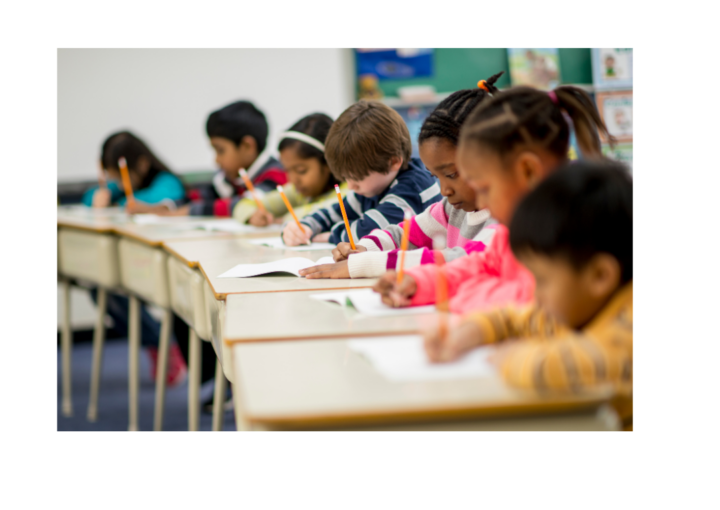 Elementary school students seated at classroom desks, writing in notebooks and completing schoolwork.
